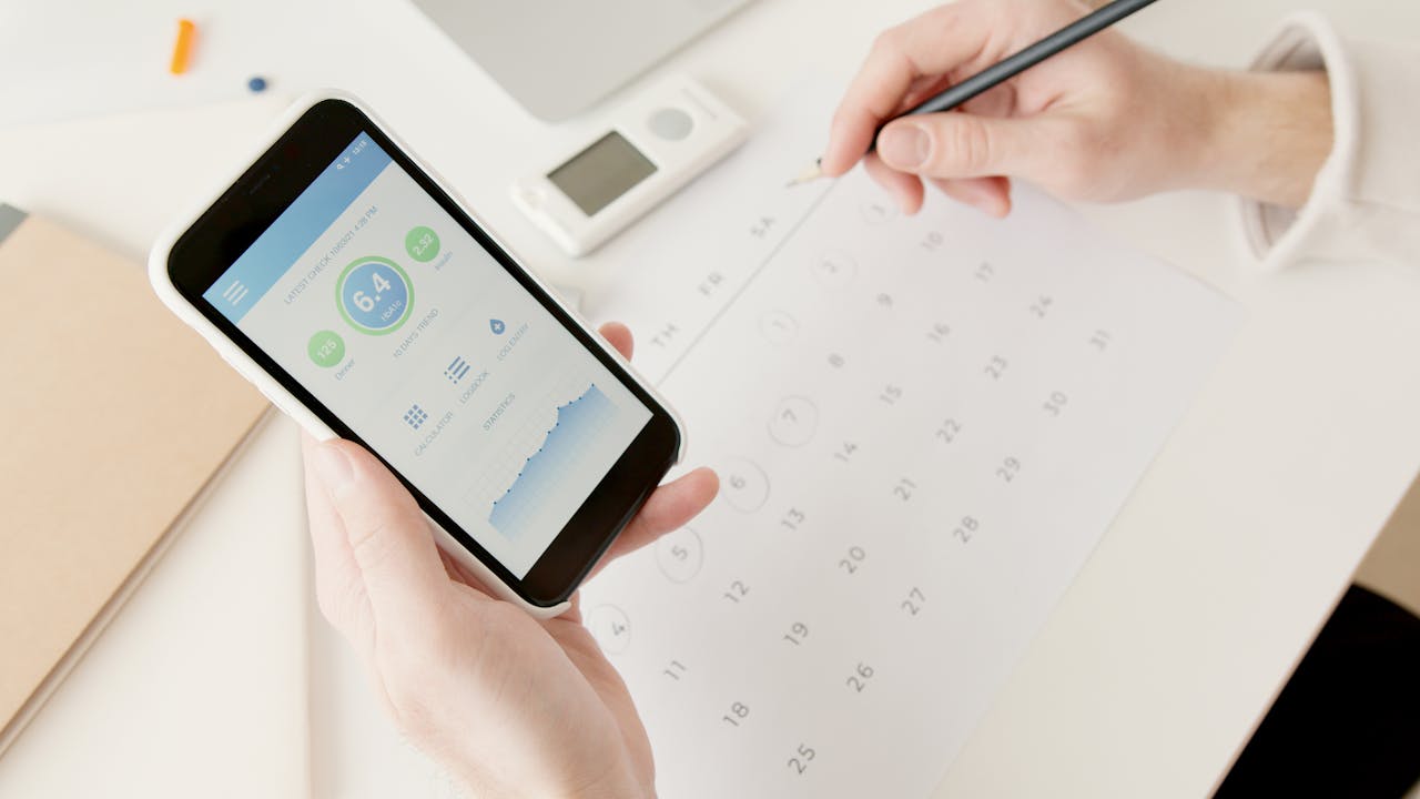 Close-up of hands using a smartphone to track health stats while planning on a calendar.